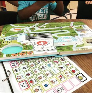 picture of core vocabulary communication board and open "Busy Helicopter book" with prek student in background