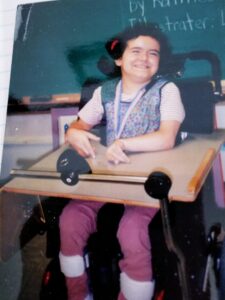 young girl with brown hair and a big smile, sitting in a wheelchair with a head switch, tray and mount for her communication device on her chair as well. 
